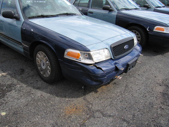 Front corner of a blue and light blue Ford Crown Victoria with a damaged bumper, parked next to another similar vehicle