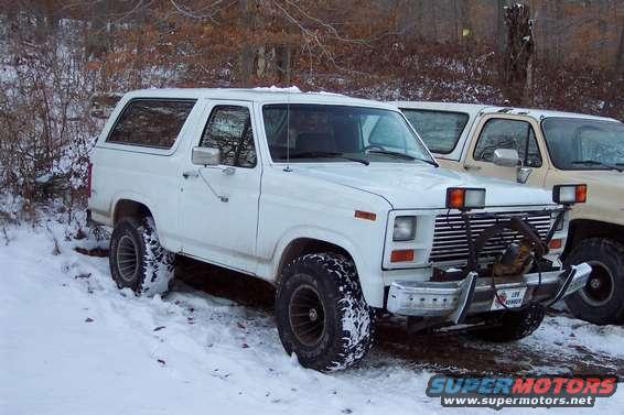 White 1986 Ford Bronco in snow with plow attached to front bumper. Another vehicle is parked behind it