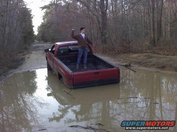 Red pickup truck stuck in muddy water, viewed from rear. A person stands in the truck bed