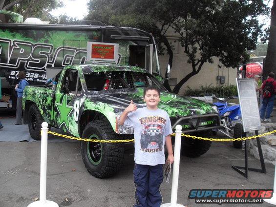 Boy gives thumbs up in front of a custom green and black off-road truck with 'ARMY' on the side. A blue ATV is parked behind it
