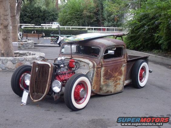 Rusty pickup truck with a surfboard on top, red engine, and whitewall tires