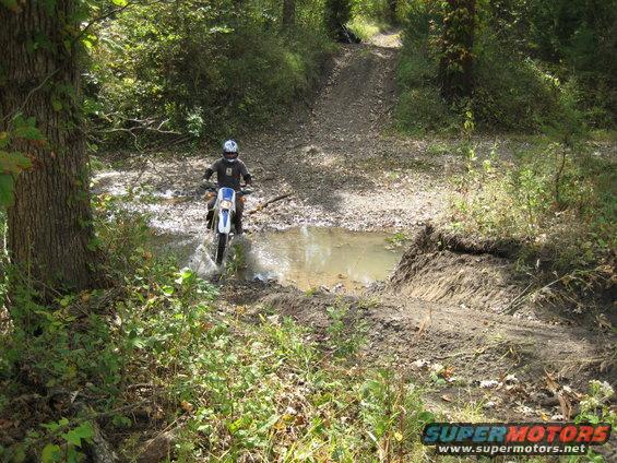 Dirt bike rider splashing through a muddy puddle on a wooded trail