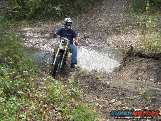 Dirt bike rider wearing a helmet and goggles splashes through a muddy puddle on a wooded trail