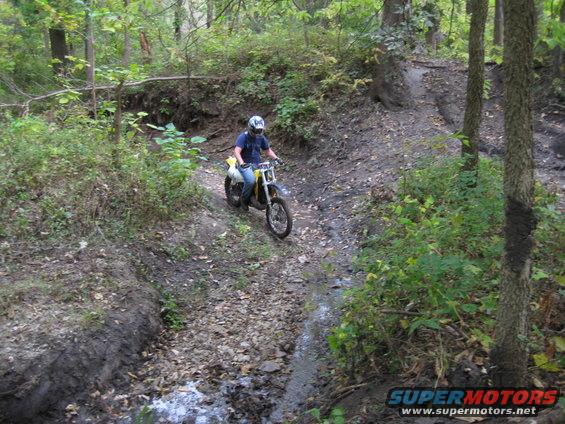 Dirt bike with yellow plastic and a rider in a blue shirt and white helmet navigates a muddy, wooded trail