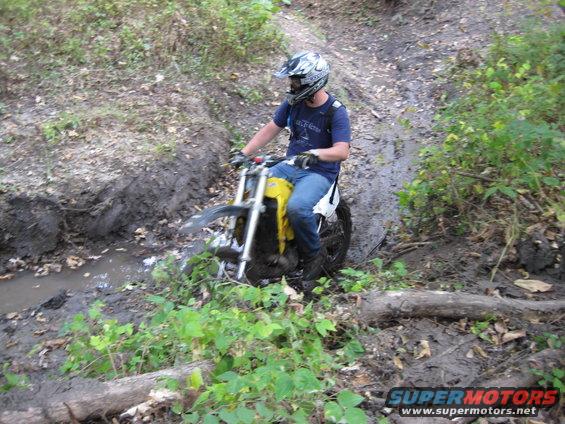 Dirt bike with yellow tank navigating a muddy trail with dense foliage