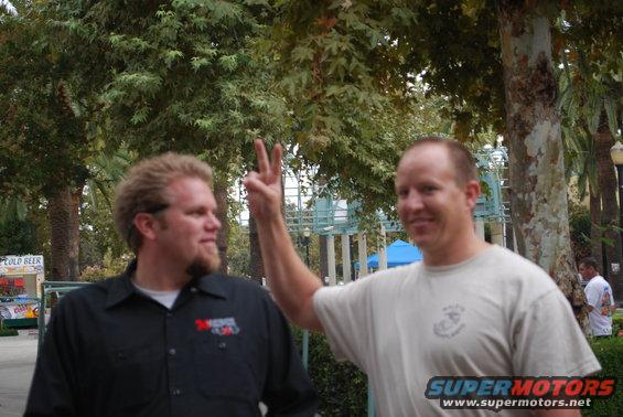 Two men stand outdoors with trees and a structure in the background; one man wears a black shirt with logos, the other a light-colored shirt