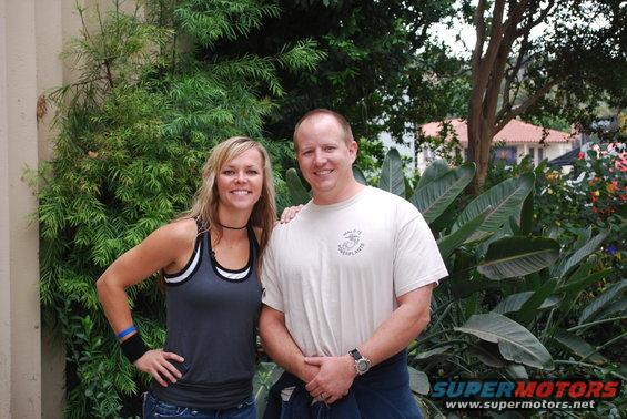 Couple posing in front of plants with a building in the background. "SUPER MOTORS" logo visible
