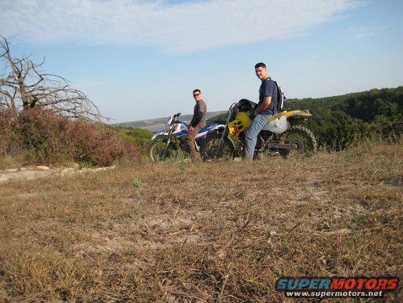 Two dirt bikes parked on a hill with riders. Dry grass and bushes surround them