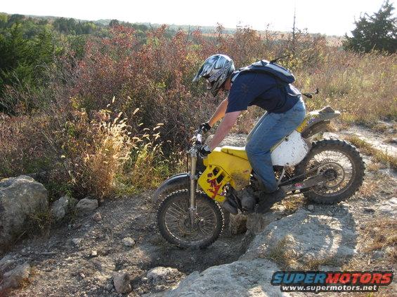 Man on yellow dirt bike navigating rocky terrain with dry brush and trees in background