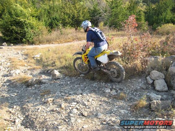 Rider on yellow dirt bike navigating a rocky, off-road trail with trees in the background