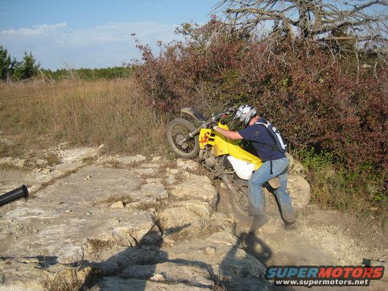 Guy struggling to push a yellow dirt bike up a rocky trail. He's wearing jeans and a helmet