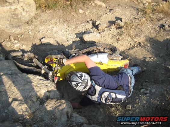 Yellow 1993 Suzuki RMX250 dirt bike on its side in rocky terrain with a rider partially underneath