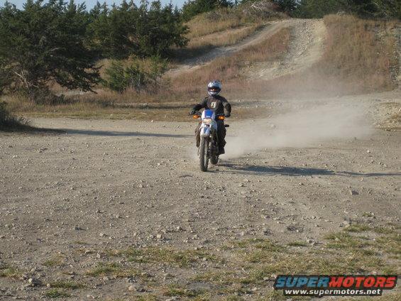 Rider on dirt bike kicking up dust on a gravel path with hills and trees in background