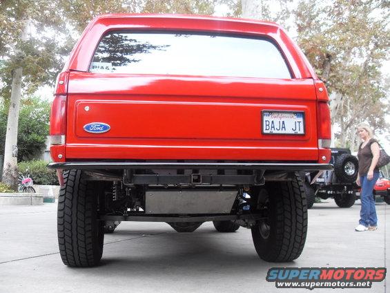 Rear view of a red 1990 Ford Bronco with a California license plate reading BAJA JT. Another vehicle is visible to the right