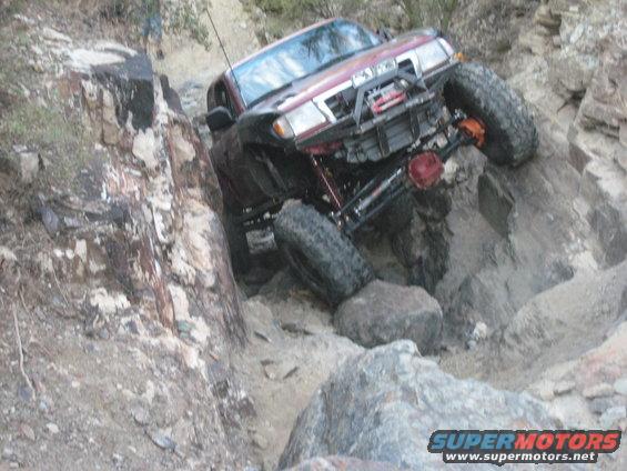 Red SUV with large tires navigating a steep, rocky incline. Visible text: SUPERMOTORS.NET