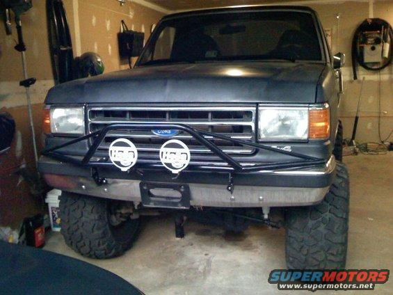 Front view of a lifted, dark gray Ford F-Series truck with a brush guard and Hella lights. It's in a garage