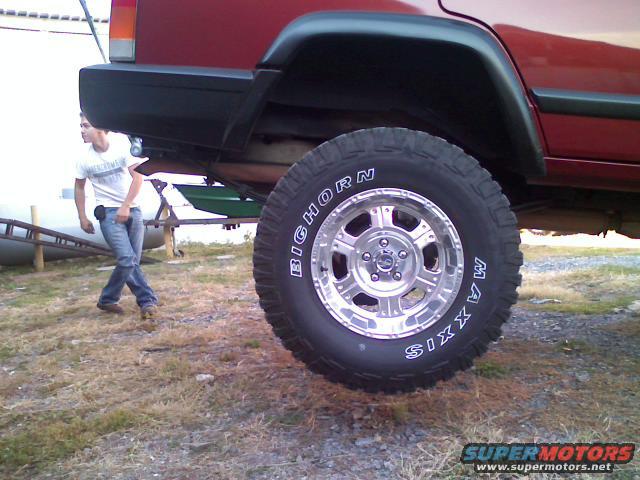 Red 1998 Jeep Cherokee rear quarter panel and large off-road tire, with a person walking in the background