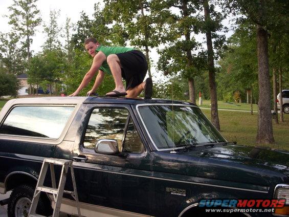 Man climbing on the roof of a dark green 1993 Ford Bronco with a ladder nearby
