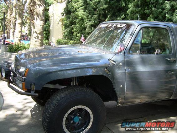 Gray pickup truck with off-road tires and a custom bumper. It has a 'NITTO' sticker on the windshield and American flags on the front fenders