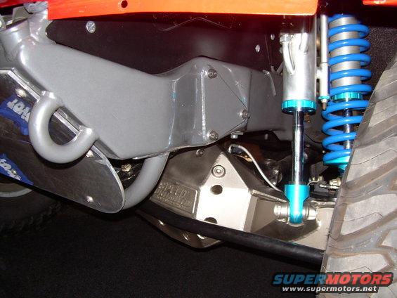 Close-up of off-road vehicle suspension with blue coilover shock and skid plate. Text on skid plate is partially visible