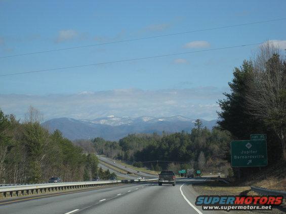Highway driving past trees towards mountains under a blue sky with clouds