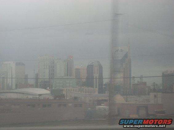 City skyline with tall buildings under a hazy sky, with a Supermotors logo in the lower right corner