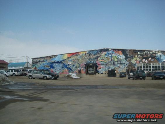 Building with a large, colorful mural on the side, parked cars in front, clear blue sky