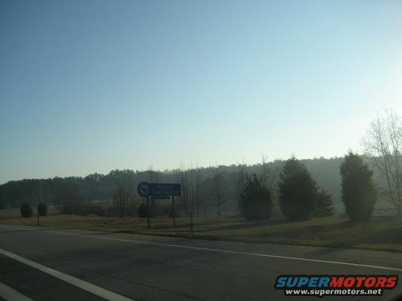 Roadside view with trees and a blue sign indicating a town name