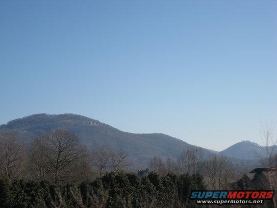 Mountain landscape with trees and houses under a clear blue sky. Watermark reads SUPERMOTORS