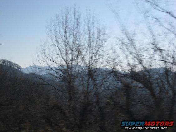 View from a moving vehicle shows bare trees and distant mountains under a hazy sky. Supermotors logo in the corner