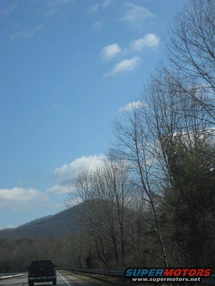 Truck driving on a highway with a wooded hillside in the background under a blue sky