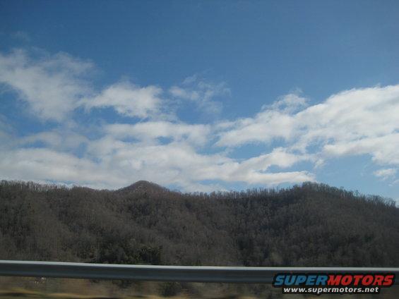 Metal guardrail in foreground with bare trees on a hill under a blue sky with clouds