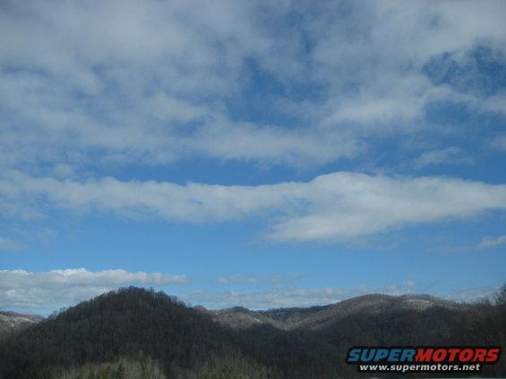 Blue sky with scattered clouds over dark, tree-covered mountains