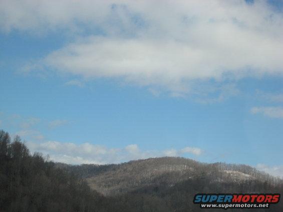 Hilly landscape with bare trees under a blue sky with scattered clouds. "SUPERMOTORS www.supermotors.net" logo in lower right