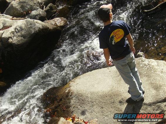 Person in Bass Pro Shops t-shirt and jeans stands on rocks next to a flowing stream