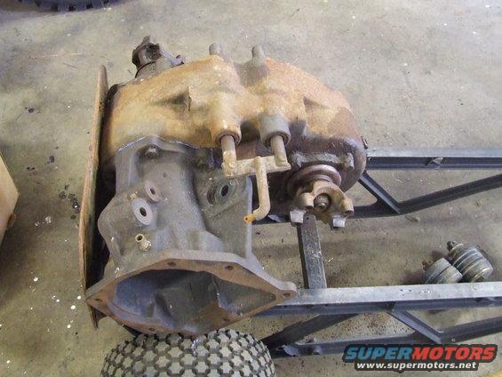 Rusty transfer case with PTO cover removed, mounted on a metal frame with a tire underneath