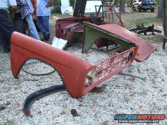 Red Ford Bronco fender and grille assembly disassembled on gravel. Bronco script visible on fender