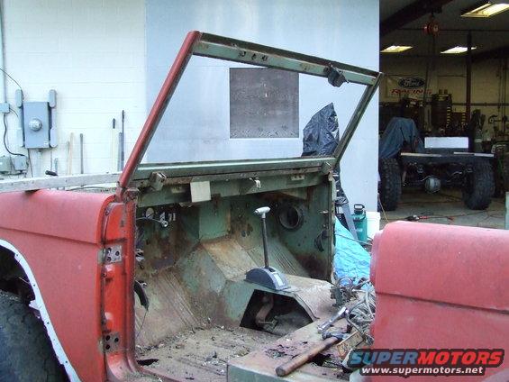 Red Ford Bronco body with no doors or roof, showing rusted interior and floor. Another vehicle chassis is visible in the background