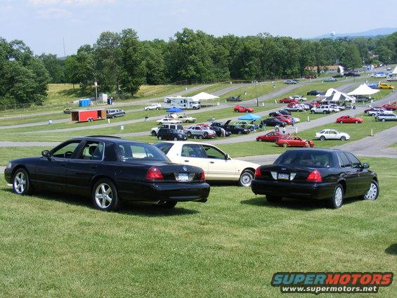 Two black 1997 Mercury Grand Marquis sedans parked on grass with other cars and tents in background