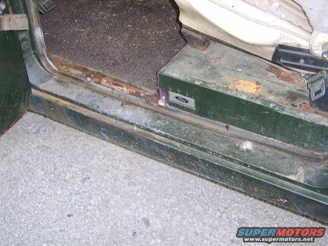 Close-up of a vehicle's rusty door sill and floor mat, with a Ford emblem visible on a green metal panel