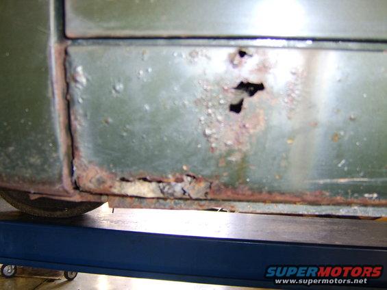 Close-up of heavily rusted rocker panel on a green vehicle, showing holes and corrosion