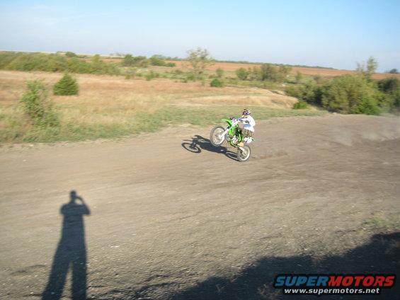 Dirt bike rider performing a wheelie on a dirt track with a dry, grassy landscape in the background