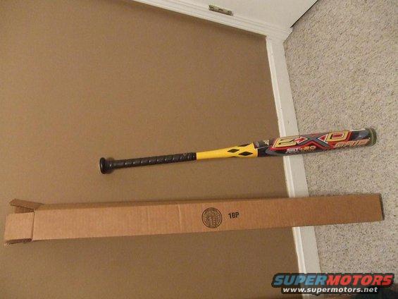 Baseball bat with yellow and black accents lies next to a long cardboard box on a carpeted floor