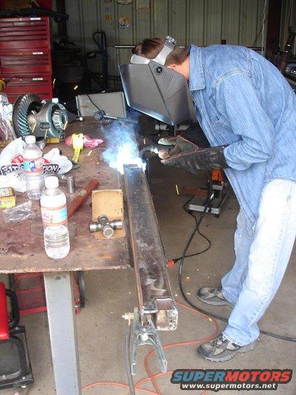 Welder in blue shirt and jeans welding a metal beam on a workbench with a differential in the background