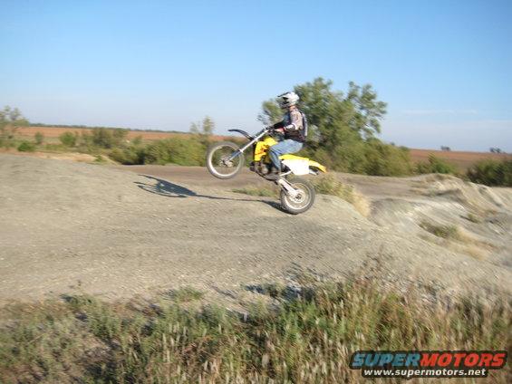Motorcyclist performing a wheelie on a dirt track with dry grass and trees in the background