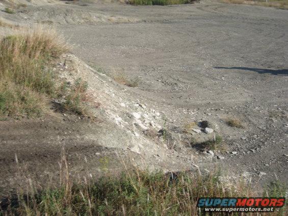 Dirt track with tire marks and a grassy embankment, dry vegetation in foreground