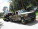 1989 Ford Bronco - A Ford Bronco is transported on a trailer pulled by a camouflage Ford F-Series truck, with a white van visible in the background.