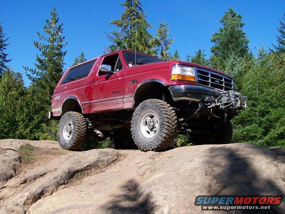 Red Ford Bronco with lifted suspension and large tires climbing over rocks on a trail