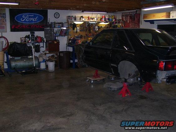 Black coupe on jack stands in a garage, missing front fender and rear bumper