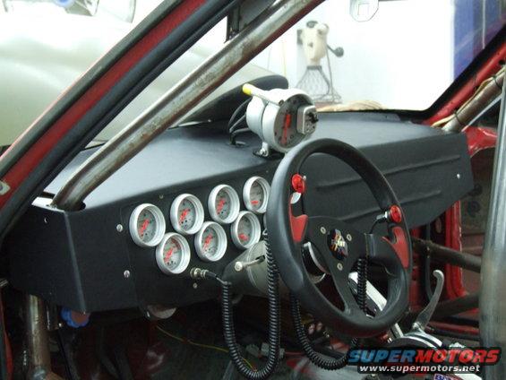 Racing car interior with multiple gauges, a black and red steering wheel, and roll cage